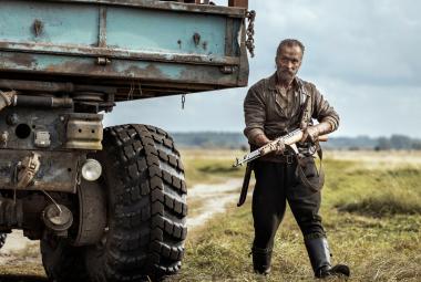 Kuvassa on vasemmalla kuorma-auton korkea takaosa.  Sen vieressä seisoo Jorma Tommila ase kädessään vihreässä maastopuvussa. Taustalla on vihreää ruohoa ja hiekkatietä. 