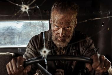 Kuvassa on Jorma Tommila ajamassa sotilasajoneuvoa ja tuulilasissa on luodin reikä.  Autossa on tummat seinät ja takaa näkyy avoin ikkuna.