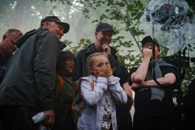 Kuvassa on Siiri Virtanen keskellä vaalean sinisessä takissa. Ympärillä on aikuisia miehiä ja naisia tummissa takissa ulkona lehtipuiden alla. 