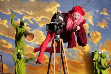 Kuvassa taustalla kelta-harmaita pilviä sinisellä taivaalla. Vasemmalla on kaksi vihreätä hiireksi pukeutunutta hahmo ja toinen seisoo okapäillä. Keskellä on vanhanaikainen filmikamera ja siinä päällä roikkuu nainen punaisissa saappaissa legginsseissä sekä viininpunaisessa takissa. Takana on pieninä kaksi vihreätä hiirtä.