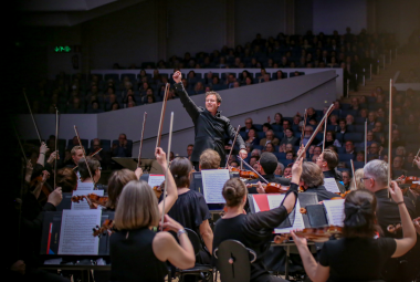 Kuvassa on keskellä Matther Halls johtamassa Tampereen Filharmoniaa.  Orkesterin soittajat ovat selin. Taustalla on osittainn tummaa.