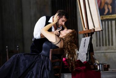 Kuvassa on Toscan roolin laulaja tumman sinisessä pitkässä puvussa istumassa tuolissa ja nojaa selkänojaan.  Hänen ylläpuolellaan on kuvartuneena kasvoja kohti mies valkoisessa puserossa ja mustassa liivissä. Tausta on tumma.