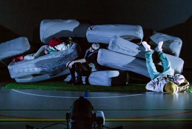 Kuvassa on isojen harmaiden tyynyjen kasa ja välissä on kaksi näyttelijää. Yksi on jalat ylöspäin makaamassa lattilalla oikeassa kulmassa.