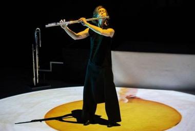 Kuvassa on Claire Chaise soittamassa huilua seisten ja hänellä on musta hihaton musta mekko sekä pitkät housut. Hän seisoo keltaisen, pyöreän maton päällä ja sen ympärillä on valkoinen ympärillä. Taustalla on mustaa.