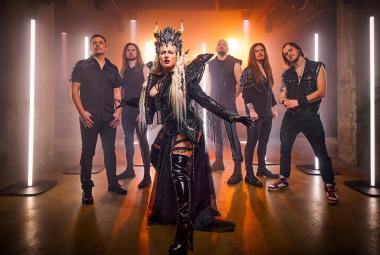 Kuvassa on edessä yhtyeen naissolisti mustassa puvussa, jossa on siipimäiset olkapäät ja päässä kruunu. Taustalla on viisi yhtyeen miesjäsentä seisomassa mustissa vaatteissa. Taustalla on pystyledvaloja ja seinien värit violettia ja oranssia.