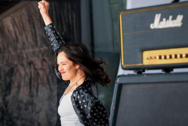 Kuvassa on Oona Airola, joka on lavalla oikea käsi pystyssä valkoisessa puserossa sekä mustassa jakussa. Taustalla näkyy tummaa seinää oikealla on kaiuttimen yläosassa tekstiä ja alemana mustaa kaiutinta. 