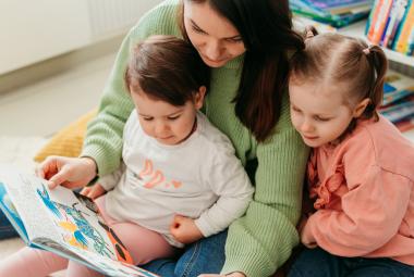 Kuvassa on nuori nainen, joka lukee kirjaa kahdelle sylissään olevalle lapselle. He ovat noin neljä- ja kuusivuotiaita. 