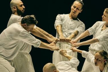 Kuvassa on viisi miestanssijaa ja yksi näinen vaaleissa kangasasuissa ja he pitävät kahdella kädellä kiinni vaaleasta renkaasta. Edessä oleva tanssija on alhaalla kumartuneena.