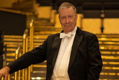 Kuvassa on Okko Kamu seisomassa portaikossa, jossa on metalliset kaiteet. Hänellä on frakki päällään ja pitää oikeata kättään kaiteella. Kuvan sävy on ruskehtava.