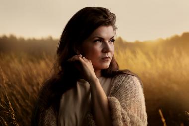 Kuvassa on Suvi Teräsniska puolivartalokuvassa ja hän katsoo oikealle. Taustalla on auringonlaskun kellertävää ja viljapelto, jossa on usvaa.