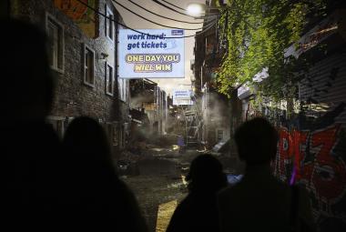 Kuvassa on tuma katunäkymä ja siinä savua ja rikkonaisia telineitä sekä kyltti, jossa lukee One day you will win.   