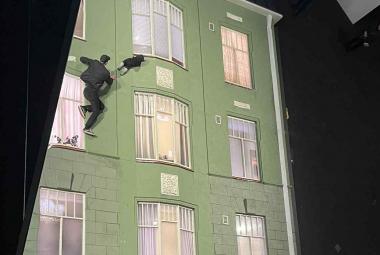 Kuvassa on molemmin puolin mustaa ja keskellä on vaalean vihreä vanha kerrostalo, jossa on kolmeen osaan jakautuneet ikkunoita kolmessa rivissä. Vasemmalla ylhäällä on nuori mies mustassa puvussa mukanaan vauva kiipeämässä seinällä.