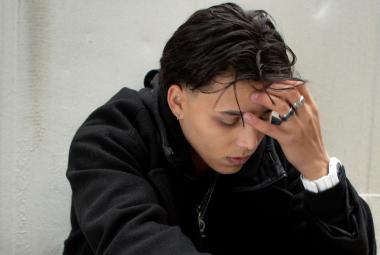 Kuvassa on Averagekidluke istumassa puolivartalokuvassa kuvartuneena. Hän pitää vasemmalla kädellään otsaansa. Tausta on vaalean harmaa.