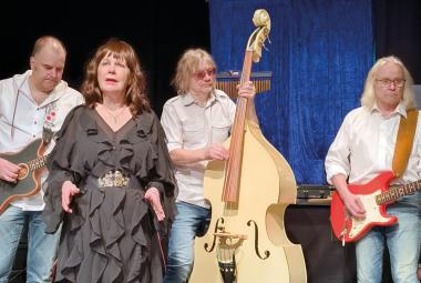 Kuvassa on vasemmalta kitaristi ja hänen vieressään edessään nainen ruskeassa rimpsutetussa mekossa ja keskellä on vaaleaa bassoa soittava mies. Oikealla on kitaraa soittava mies valkoisessa paidassa ja farkuissa.