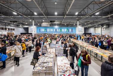 Kuvassa on isossa hallissa useita pitkiä pöytiä täynnä kirjoja ja niitä katselemassa on paljon ihmisiä. Takana on iso sininen lakana, jossa on valkoisin kirjaimin Ilmaiskirjapäivä.