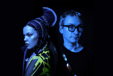 Kuvassa on puolivartalokuvassa vasemmalla Hilda Länsman sivukuvassa ja hänellä on Lapin puvun päähine ja puku. Oikealla on Tuomas Norvio mustassa t-paidassa. Kuvan sävy on tumma.