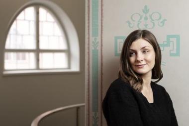 Kuvassa on oikealla Iida Turpeinen puolivartalokuvassa mustassa puserossa. Hän on tilassa, jossa on vaalea, turkoosikoristeinen seinämän reunus. Taaempana näkyy kaarevaa ikkunaa.