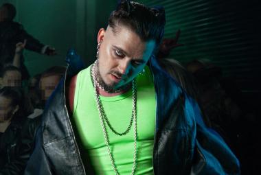 Kuvassa on Käärijä puolivartalokuvassa. Hänellä on hihaton vaalean vihreä t-paita ja musta nahkatakki olkapäillään kevyesti. Taustalla on tummahkoa ja alempana näkyy yleisön kasvoja.