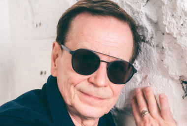 Kuvassa on Kake Randelinin kasvot.  Hänellä on aurinkolasit päässään ja hän pitää oikeaa kättään vaaleassa tiilisessä seinässä.  