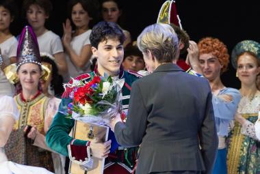 Kuvassa on keskellä tummahiuksinen Giulio Diligentte puolivartalokuvassa vihreässä roolipuvussa ja hänellä on kunniakirja oikeassa kädessään. Hänelle on ojentamassa kukkakimppua nainen, jolla on harmaa jakku ja hän on selin. Taustalla on Kansallisbaletin tanssijoita kolmessa rivissä rooliasuissaan. 