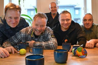 Kuvassa ovat yhtyeen viisi jäsentä vaalean ruskean pöydän ääressä istumassa eri värisissä paidoissa. Edessä on sinisiä mukeja.