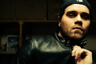 Kuvassa on oikeassa reunassa puolivartalokuvassa ja hänestä näkyy mustalla pohjalla kasvot ja vasenta kämmentä. Hänellä on musta nahkatakki päällään.