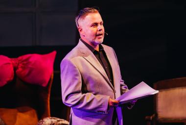 Kuvassa on keskellä Pekka Laasonen seisomassa teatterin penkkien välissä ja seisoo sivuttain. Hänellä on vaalea takki päällään ja käsikirjoitus kädessään. Tausta on tumma.