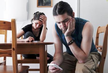 Kuvassa on etualalla oikealla nuori mies istumassa, nojaa vasemmalla kädellään poskeensa ja katsoo oikeassa kädessään olevaa kännykkää. Pöydän takana istuu nuori nainen ja katsoo myös kännykkäänsä. 