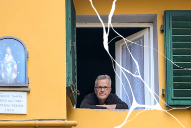 Kuvassa on Olli Kortekangas avatun ikkunan alareunassa puolivartalokuvassa. Hänellä on m usta paita päällään.  Talon seinä on keltainen ja vasemmalla on vaalean sininen madonnan ja Jeesus-lapsen kuva. Oikealla on toinen vihreä ikkunaluukku.