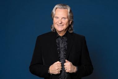 Kuvassa on keskellä Pepe Willberg ja hänellä on musta takki sekä musta paita. Hän pitää molemmilla ksillän kiinni takin liepeistä. Tausta on sininen. 