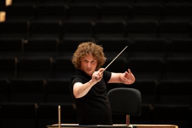 Kuvassa on Santtu-Matias Rouvali johtamassa orkesteria tahtipuikko silmiensä tasalla puolivartalokuvassa. Hänellä on musta t-paita päällään. Tausta on musta.