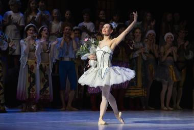 Kuvassa on keskellä valkoisessa balettipuvussa Sofia Gustafsson ja hänellä on vasen käsi ojennettuna sekä kukat oikeassa kädessään. Taustalla näkyy himmeästi muita esiintyjiä.