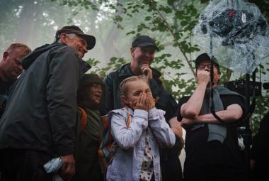 Kuvassa on keskellä vaalean sinisessä farkkutakissa Siiri Virtanen ja hänen ympärillään on kuvausryhmän kuusi henkilöä tummissa ulkovaatteissa. Yläpuolella on vihreitä lehtipuun oksia ja kaikki seisovat puun alla.