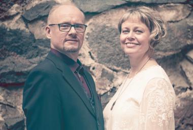 Kuvassa on vasemmalla Tapio Lappalainen  mustassa takissa ja Sanna Lappalainen vaaleassa puserossa. Taustalla on kiviseinää, jossa on suuria, tummia kiviä ja vaaleita välimuurauksia. 