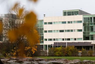  Kuvassa on vasemmalla olevan lähes lehdettomän puun takana oleva rakennus, joka on L.n muotoinen ja jonka oikea osa on vaalea. Alla näkyy litteähköjä kiviä ja pihapensaita