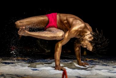 Kuvassa mies, jolla on pronssiseksi maalattu iho, ja hän käsien päällä ja muu keho vinottain vaaka-asennossa ylhäällä. Hänellä on punaiset alushousut. Tausta on musta ja lattialla on valkoista vaahtoa.