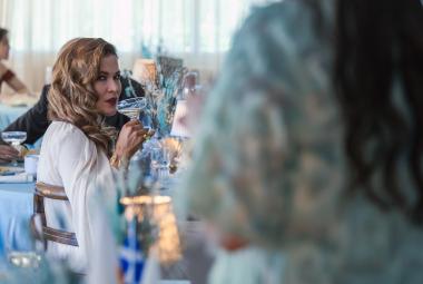 Kuvassa on ravintolatila ja oikealla edessä on himmeästi seisomassa nainen, josta näkyy turkoosin valkoista puseroa ja hieman pitkiä hiuksia selässä. Vasemmalla kauempana istuu Jenni Vartiainen sivuttain ja hän katsoo tulijaa sekä siemaileen oikeassa kädessään olevaa drinkkiä.