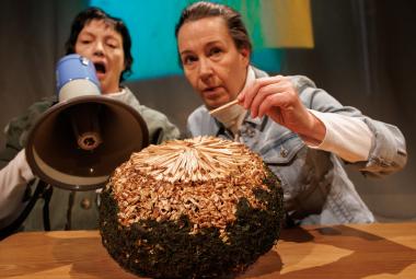 Kuvassa ovat istumassa pöydän ääressä Teija Töyry kädessään megafoni ja Heidi Syrjäkari ojentamassa vasenta kättään kohti pöydällä olevaa isoa palloa. Se on ruskea ja siinä on kultausta päällä. 