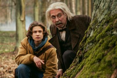 Kuvassa ovat vasemmalla metsässä kyykyssä suuren puunrungon vieressä Kuura Rossi ja hänen vieressään vähän ylempänä Martti suosalo pitkissä vaaleissa hiuksissa. Taustalla näkyy puunrunkoja.