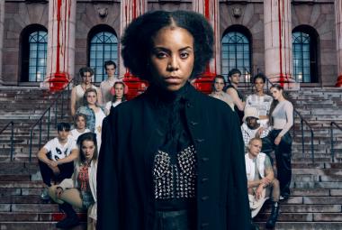 Kuvassa on keskellä mustassa mekossa Abdirahman Bile iso kokoisena ja hänen takanaan on joukko muita näyttelijäitä ryhmänä rapuilla punertavan tiilirakennuksen edessä. 