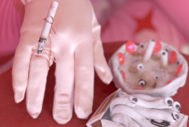 Kuvassa on pinkillä silkkisellä pohjalla käsi vaaleassa hansikkaassa ja sen päälle on kiinnitetty tupakka. Oikealla on epämääräisen muotoinen valkoinen tuhkakuppi, jossa on vaaleita keraamisia tumppeja. Edessä käyristettyjä keraamisia tupakoita.