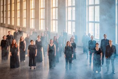 Kuvassa on Helsingin kamarikuoron jäsenet seisomassa mustissa mekoissa ja puvuissa. He ovat isossa hallissa, jossa on korkeat ruutuikkunat rivissä, joista tulee valoa.  