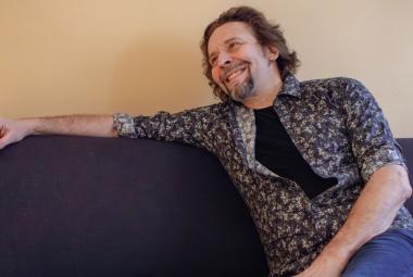 Kuvassa on Ilari Johansson istumassa mustalla sohvalla ja hänen oikea kätensä on ojentuneena sohvan selkänojan päällä. Hänellä on mustan kirjava paita päällään. Seinä on kellertävä.