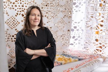 Kuvassa on vasemmalla Johanna Pöykkö mustassa mekossa puolivartalokuvassa. Taustalla on ripustettu kankaita, joissa on ruskeareunaisia reikiä erilaisina kuvioina. 