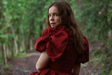 Kuvassa on Marketta Tikkanen selin kävelemässä metsätietä, jonka varrella on koivuja. Tkkasella on punainen mekko päällään ja siinä on puhvihihat.