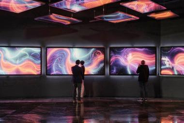 Kuvassa on keskellä ruuturivissä värikkäitä kiemurtelevia viivoja ja pintoja.  Tila on tumma ja kuvat heijastuvat kattoon ja lattiaan. Kuvia katselee pariskunta ja yksittäinen katselija.