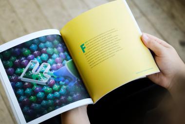 Kuvassa on kädet pitelemässä kirjaa, jossa vasen sivu on tummansininen ja siinä on kirjaintunnuksia. Oikealla keltaisella sivulla on keskellä puoli sivua tekstiä.