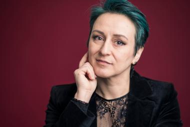 Kuvassa on Magdalena Hai puolivartalokuvassa. Hänellä on tumma pusero päällään ja hän pitää kättään oikealla poskella. Kuvan tausta on burgundin värinen.