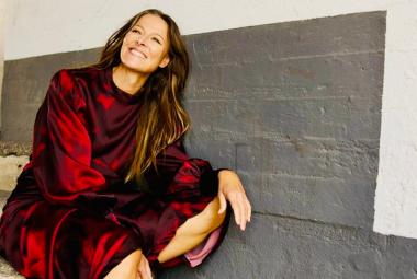 Kuvassa on Mia Hafrén istumassa betoniportailla ja nojaamassa harmaaseen betoniseinäään. Hänellä on pitkät ruskeat hiukset ja punainen mekko päällään. 