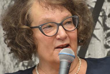 Kuvassa on Monika Fagerholm kasvokuvassa ja puhumassa mikrofoniin. Hänellä on ruskeat kiharaiset hiukset ja silmälasit.